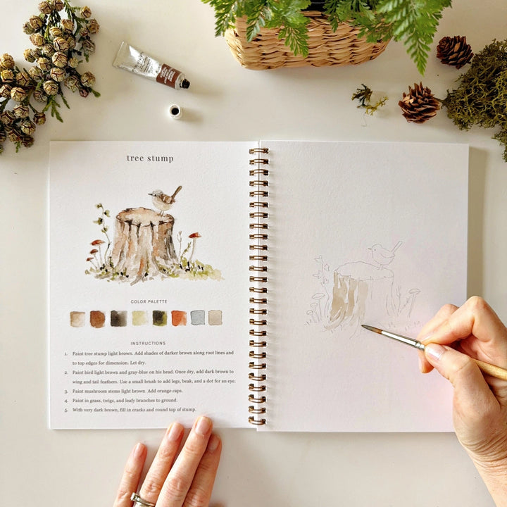 Artbook with nature-themed illustrations and color swatches, surrounded by plants and a pen.