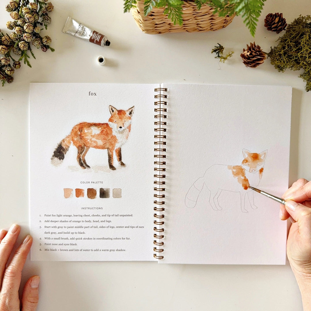 Open book with a fox illustration and color palette, being painted by a hand.