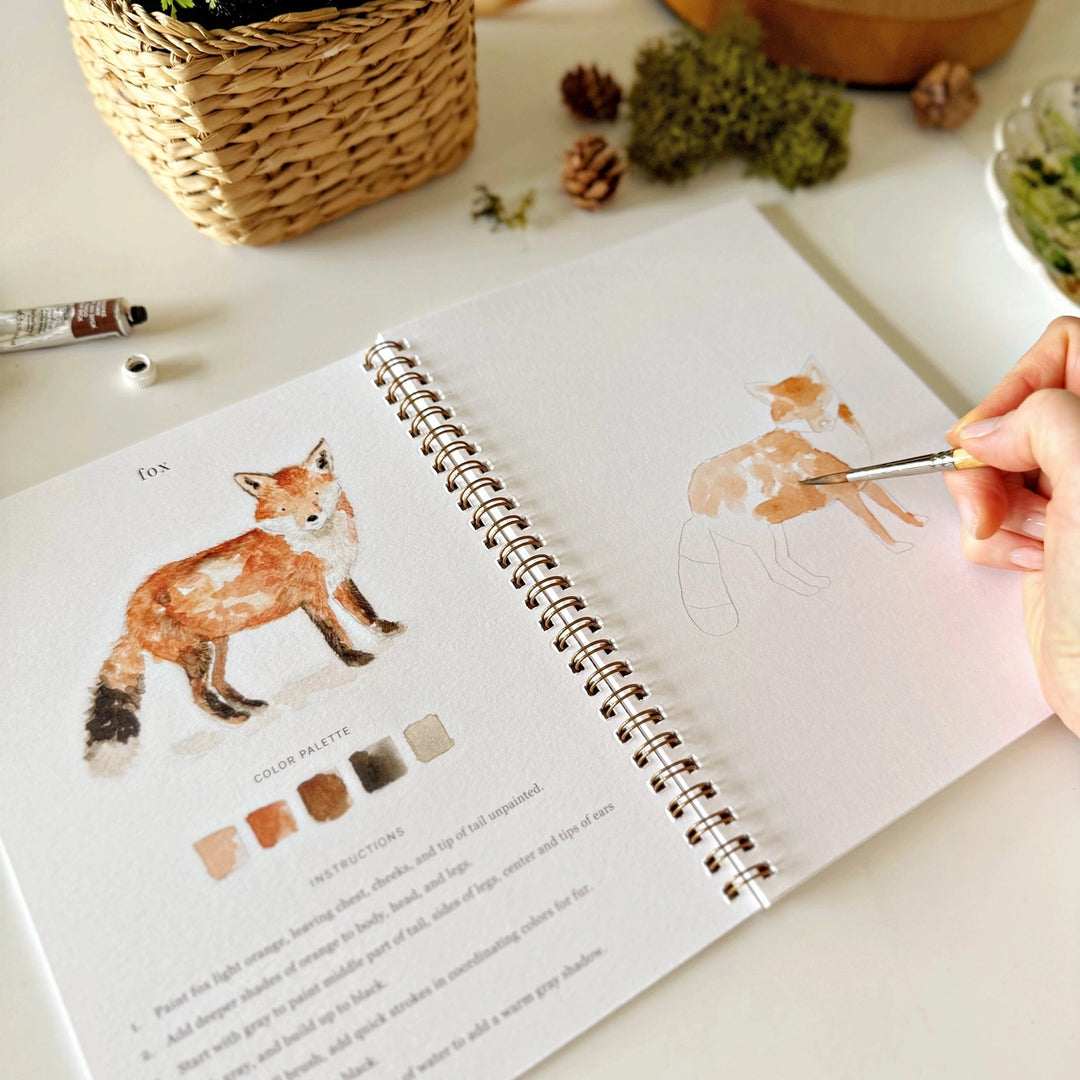 Artistic sketchbook with a fox illustration and a hand holding a paintbrush.
