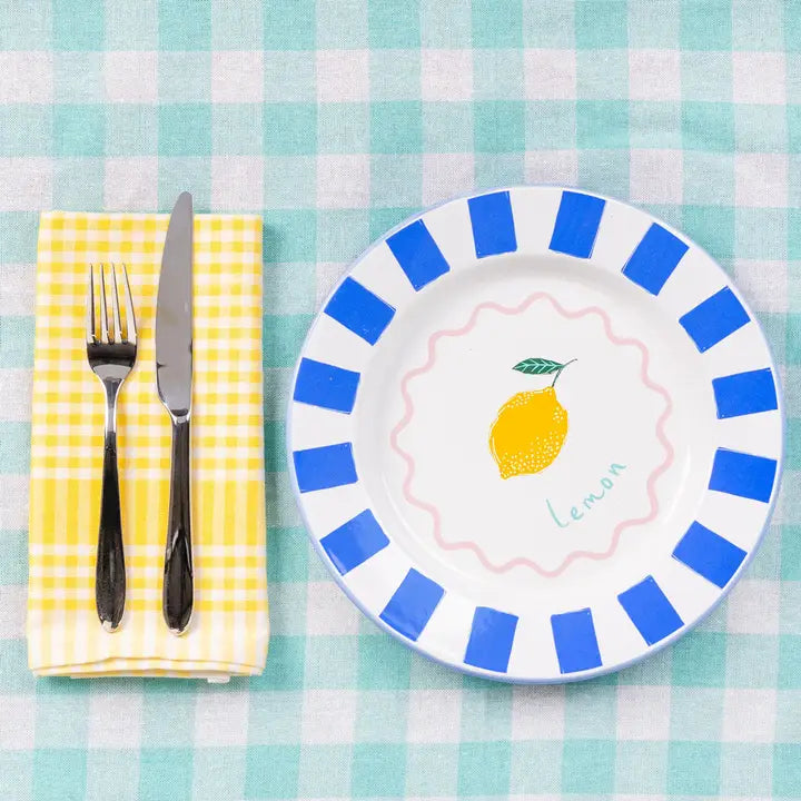 White plate with blue rim featuring a lemon design, placed on a checkered tablecloth with a fork and knife.