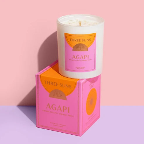 A white Three Suns AGAPI candle sits on top of its pink and orange packaging box against a soft pink and lavender studio background.