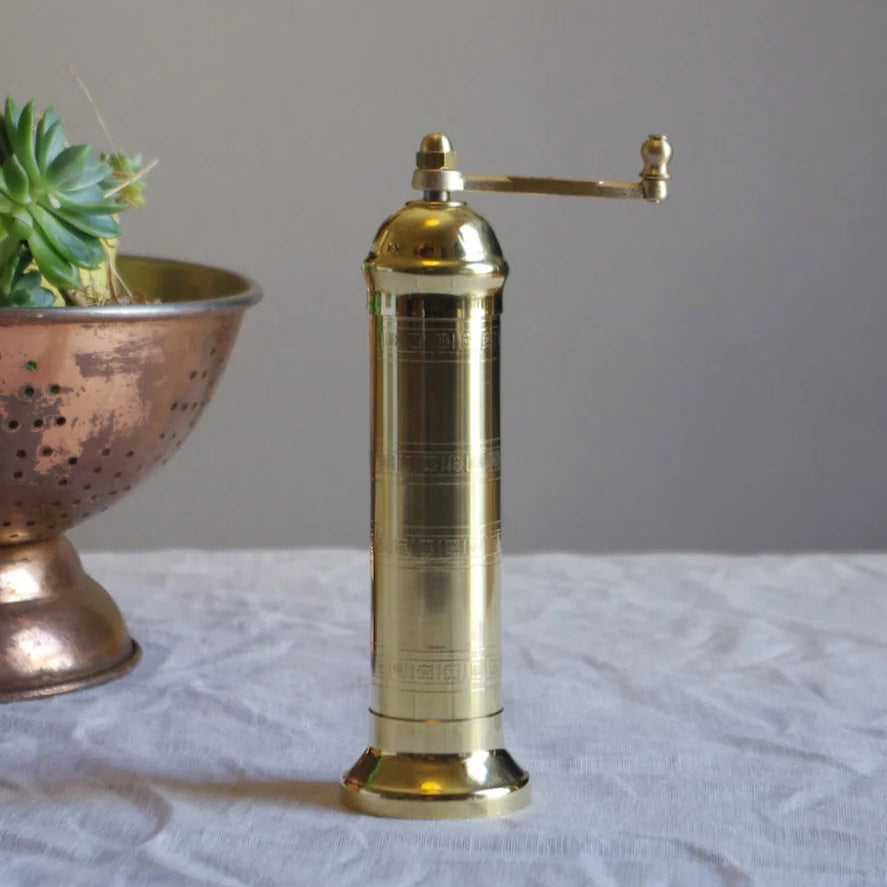 A vintage brass manual salt grinder with a horizontal handle, standing on a white fabric-covered surface with a neutral gray background.