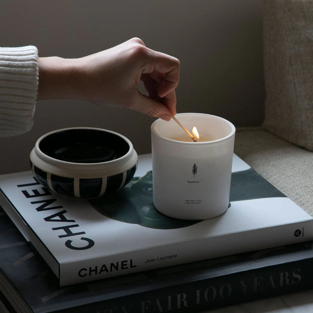 An Archipelago Aspen candle in a white vessel on a pile of books with a hand lighting the candle with a match