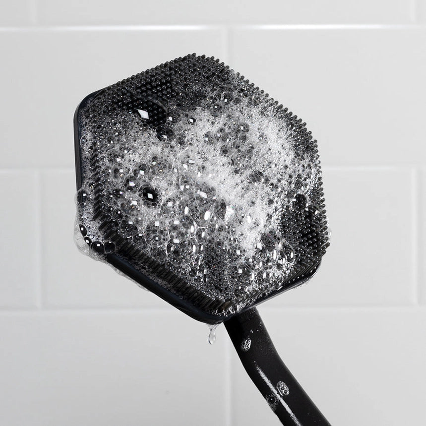 Cleaning back brush with a black handle and bristles covered in soap suds against a tiled bathroom wall.