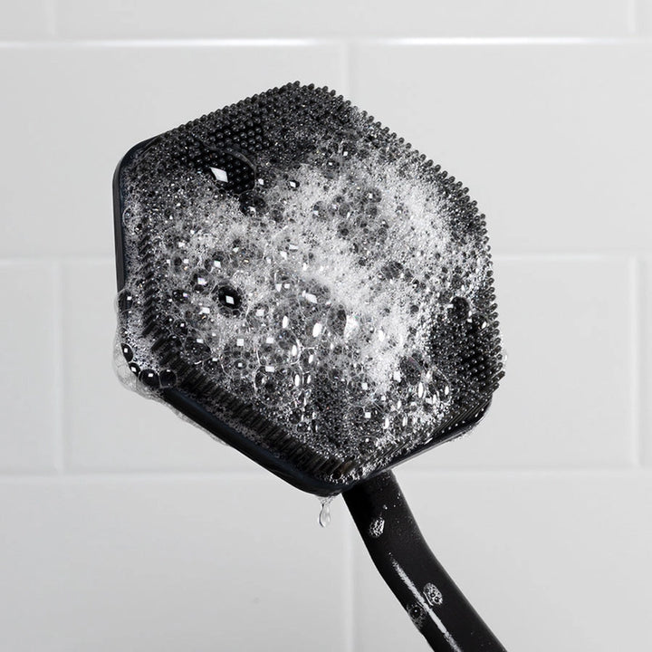 Cleaning back brush with a black handle and bristles covered in soap suds against a tiled bathroom wall.
