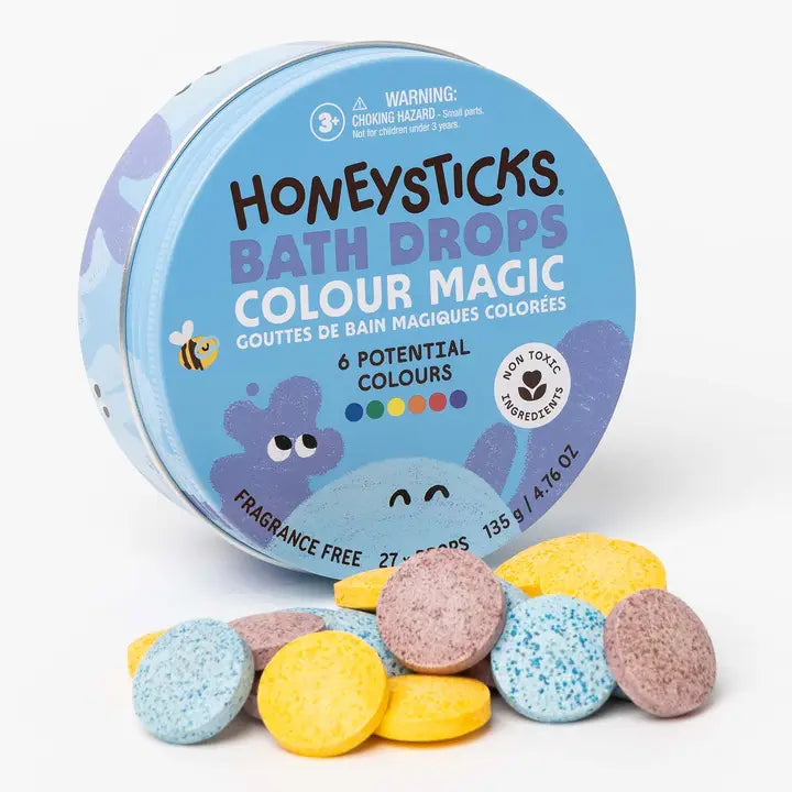 Honeysticks Bath Drops Colour Magic in a round blue tin with several loose bath drops in pastel blue, yellow, and red in front, on a white background.