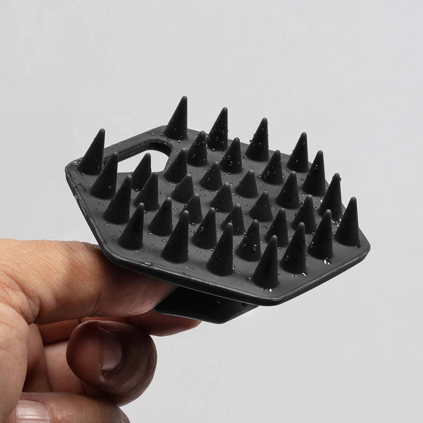 A hand holding a black silicone beard scrubber with multiple sharp conical spikes against a plain light gray background.