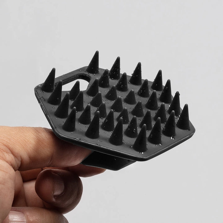 A hand holding a black silicone beard scrubber with multiple sharp conical spikes against a plain light gray background.