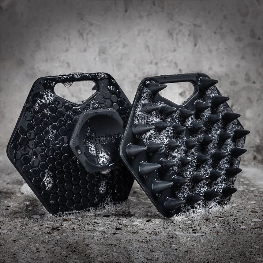 Two black silicone beard scrubbers with soap suds on a grey tile background
