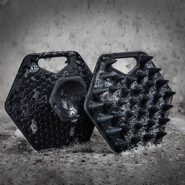 Two black silicone beard scrubbers with soap suds on a grey tile background
