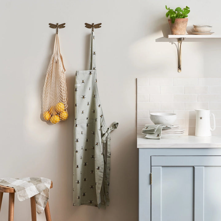 Fabric apron with small bee pattern hanging on a dragonfly shaped hook on a kitchen wall