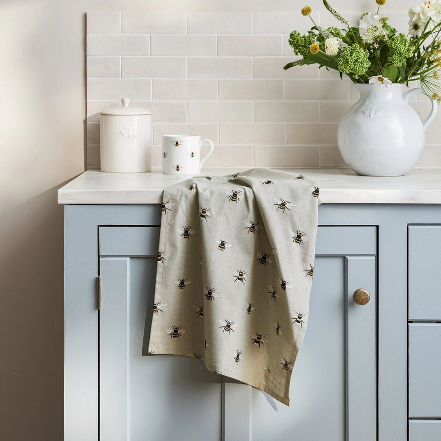 A rectangular kitchen tea towel with a hanging loop at the top left, featuring a repeating pattern of small bumblebees draped over a kitchen benchtop