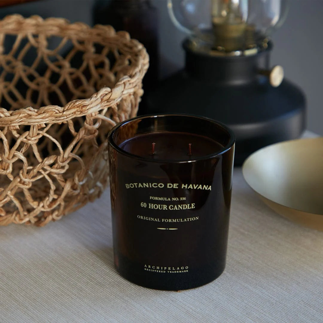 Black candle labeled 'Botanico de Havana' on a surface with a woven basket and lamp in the background.
