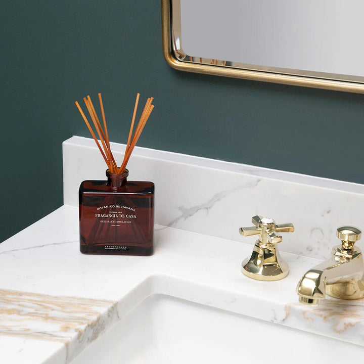 Reed diffuser on a bathroom counter with gold fixtures and a mirror.