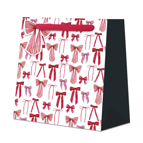 A white gift bag featuring an all-over pattern of red and pink striped and solid bow illustrations, with a red ribbon handle and a black side panel, shown on a plain white background.