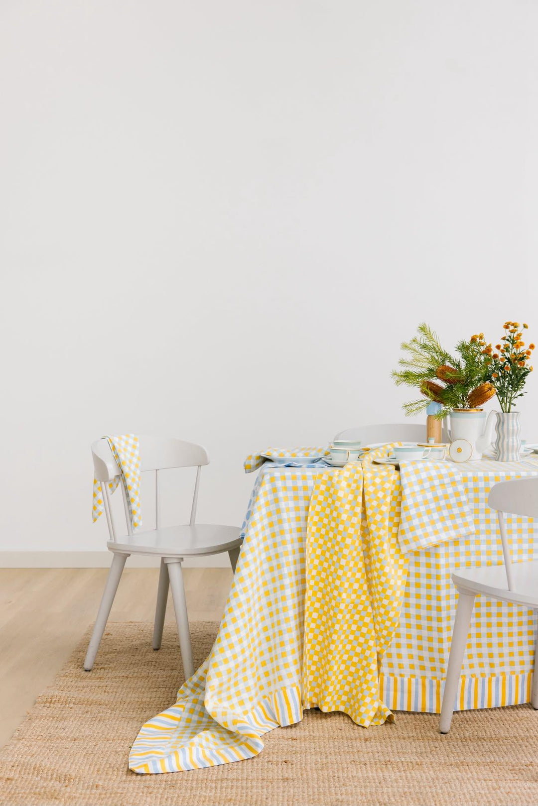 A yellow and white gingham tablecloth draped over a dining table, styled with white chairs, a vase of orange flowers, and a woven jute rug against a plain white wall.
