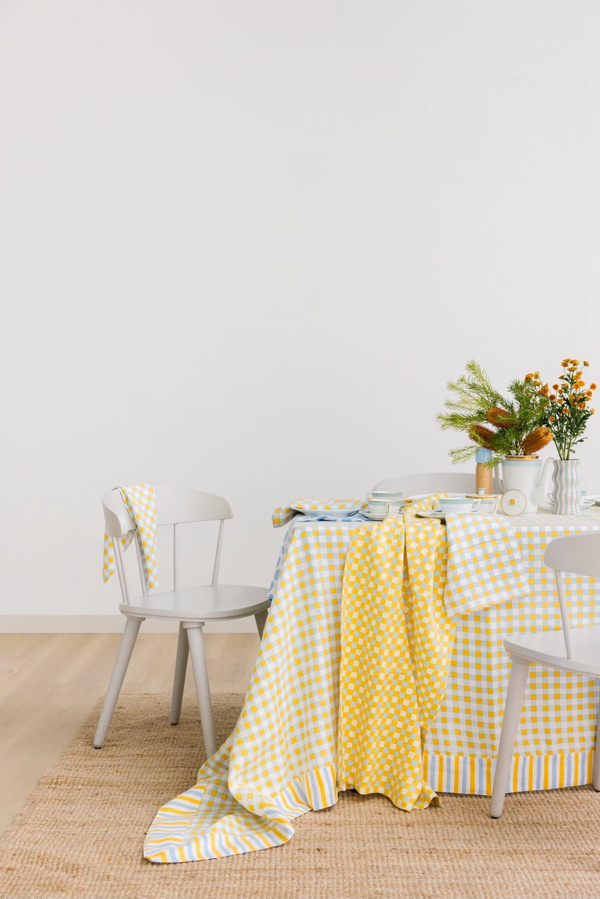 A yellow and white gingham tablecloth draped over a dining table, styled with white chairs, a vase of orange flowers, and a woven jute rug against a plain white wall.