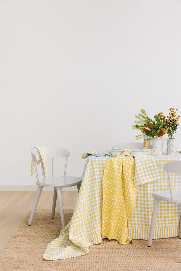 A yellow and white gingham tablecloth draped over a dining table, styled with white chairs, a vase of orange flowers, and a woven jute rug against a plain white wall.