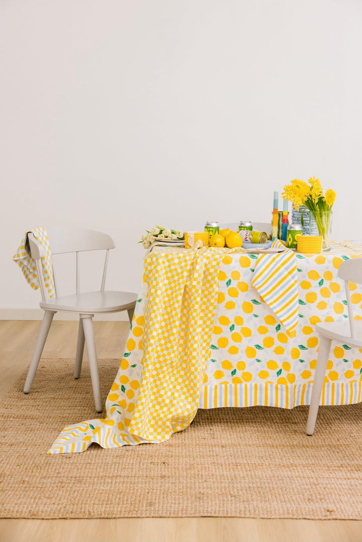 A yellow and white lemon-patterned tablecloth draped over a dining table, styled with yellow flowers, lemons, and striped and check napkins on a light wood floor against a white wall.