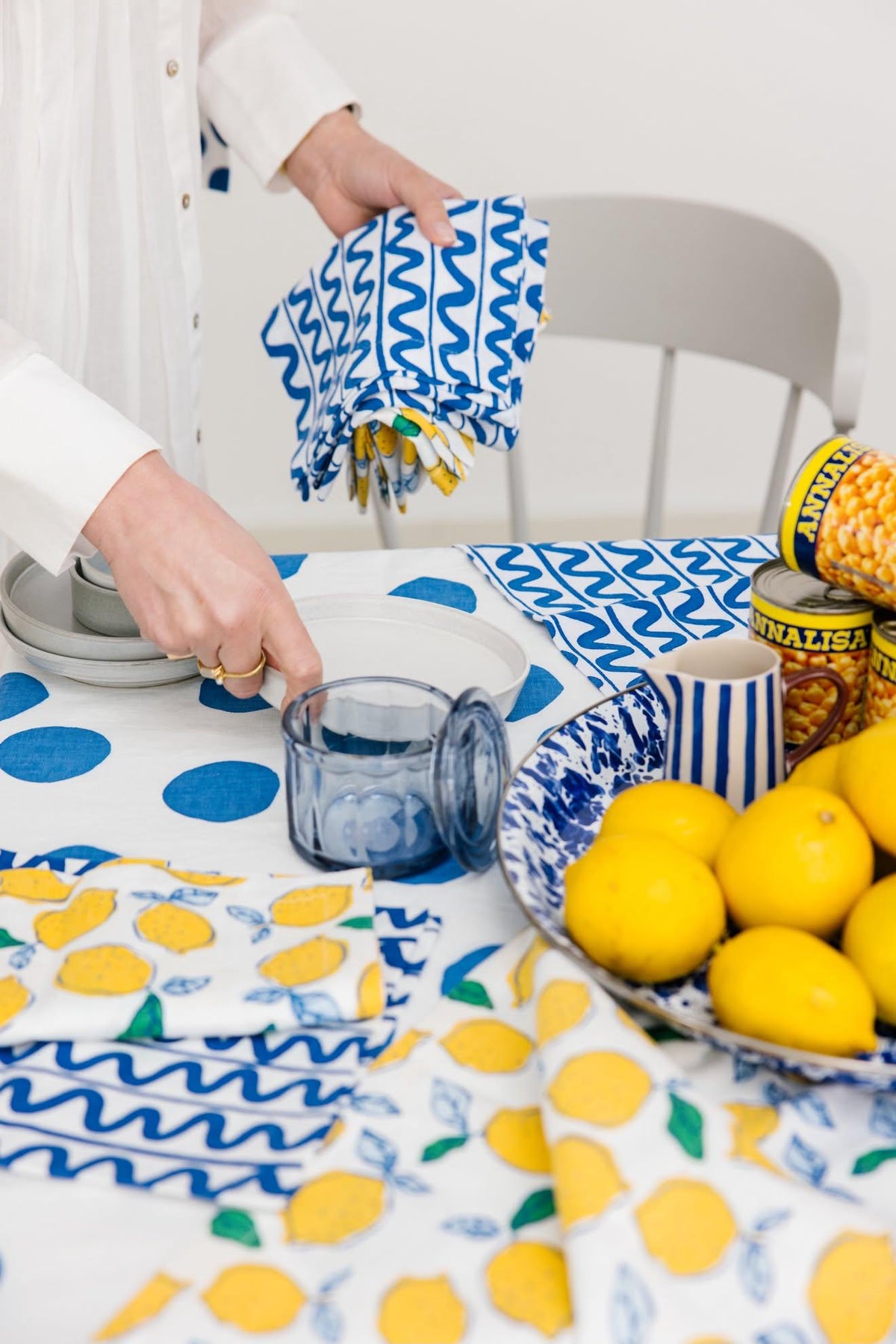 A person in a white shirt holds a blue and white patterned cloth napkin over a table styled with lemons, striped mugs, and matching patterned table linens.