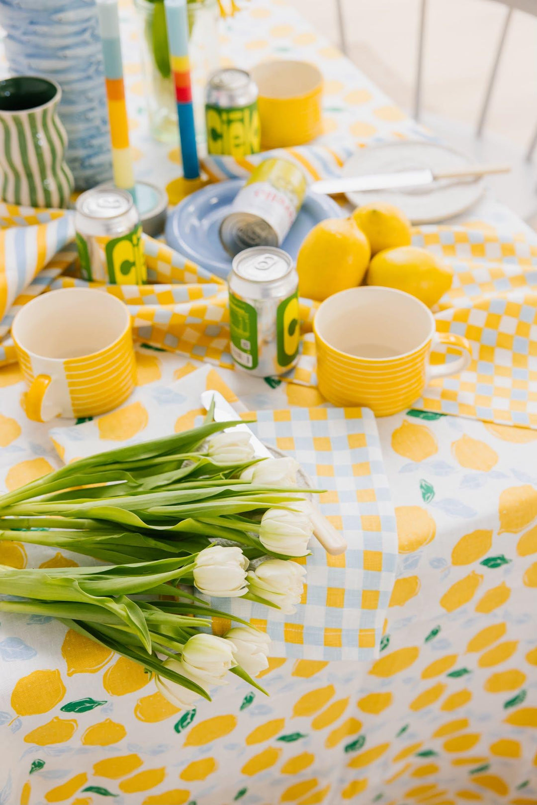 A styled table setting with a white tablecloth printed with yellow lemons and a yellow-and-white checkered pattern. Yellow mugs, lemon-themed beverage cans, whole lemons, tulip flowers, and plates are arranged on the table in a bright, airy setting.