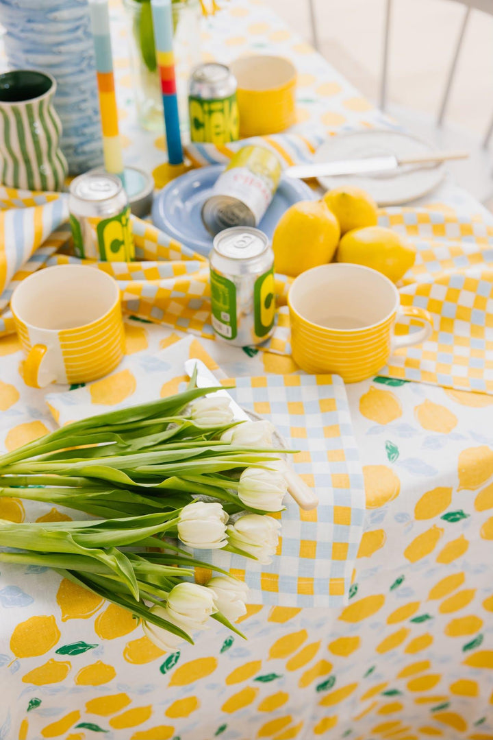 A styled table setting with a white tablecloth printed with yellow lemons and a yellow-and-white checkered pattern. Yellow mugs, lemon-themed beverage cans, whole lemons, tulip flowers, and plates are arranged on the table in a bright, airy setting.