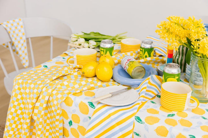 A styled dining table setup featuring a yellow and white lemon-patterned tablecloth, matching yellow and white checkered napkins, striped yellow and white mugs, yellow lemons, green and white beverage cans, white tulips, and a vase of yellow flowers.