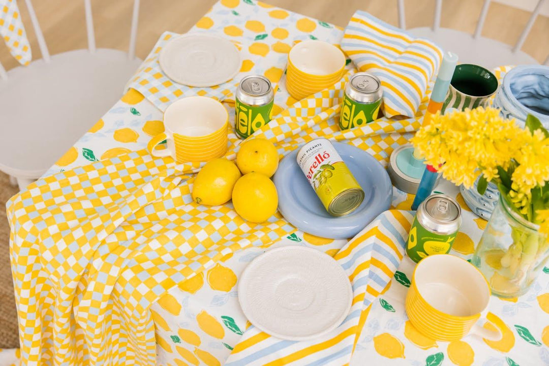 A styled picnic table setup featuring a yellow and white lemon patterned tablecloth, matching cups, plates, napkins, and soda cans, alongside lemons, a blue plate, and yellow flowers.