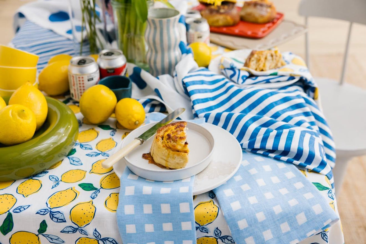 A styled outdoor dining table setting featuring a white tablecloth printed with yellow lemons, blue and white striped napkins, a green bowl of lemons, and a slice of pie on a white plate.