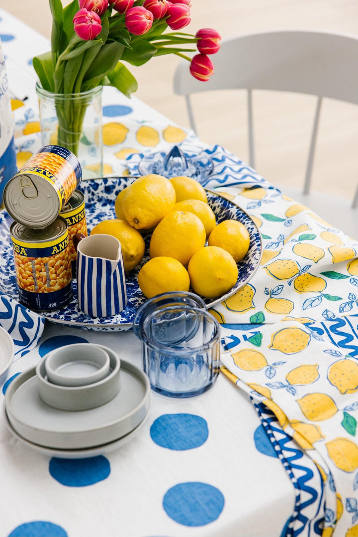 A styled dining table featuring a white tablecloth with blue polka dots and a yellow lemon print napkin, set with a blue and white patterned plate of lemons, stacked glass bowls, and a vase of pink tulips.