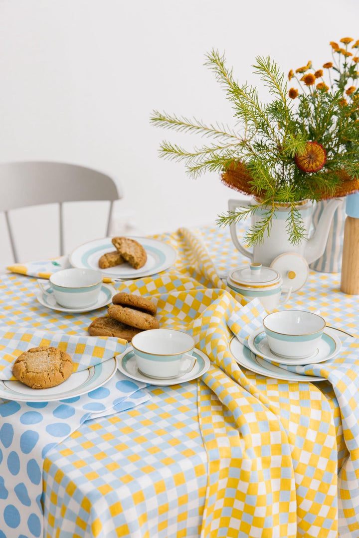 A styled dining table setting with a yellow and white checkered tablecloth, matching teacups and saucers, cookies on plates, and a floral centerpiece in a white vase against a white wall.
