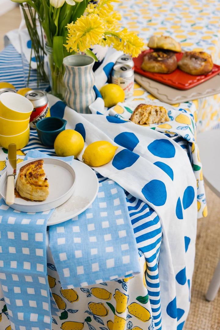 A styled dining table featuring a white tablecloth with blue polka dots and a yellow lemon print napkin, set with a blue and white patterned plate of lemons, stacked glass bowls, and a vase of pink tulips.