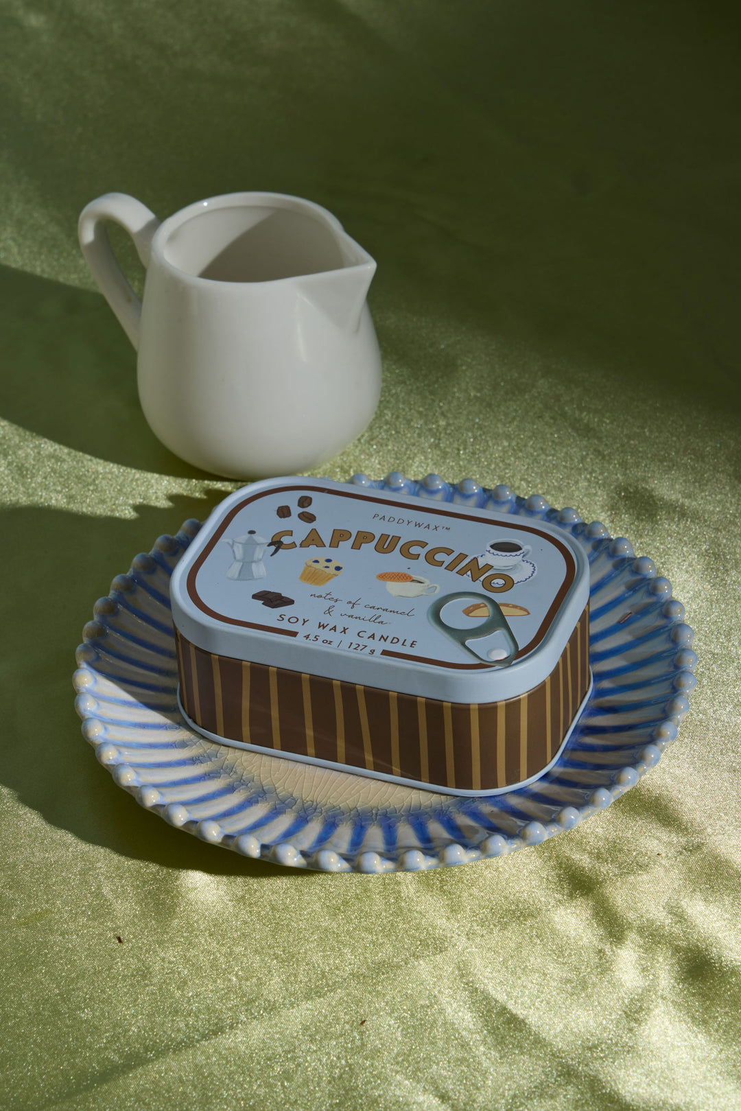 A rectangular metal candle tin labeled Cappuccino sits on a blue-and-white patterned plate, with a small white ceramic pitcher behind it on a green fabric surface.