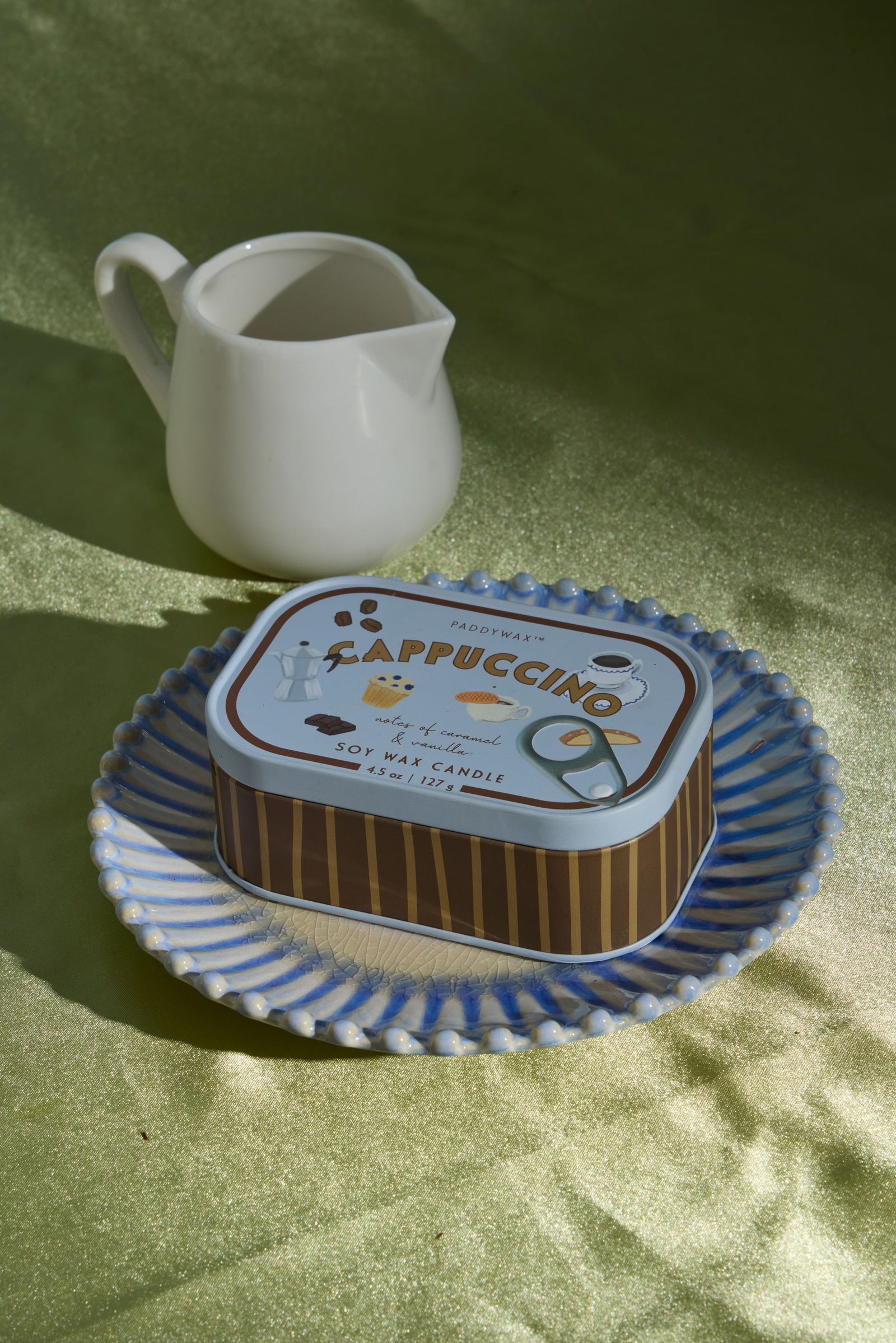 A rectangular metal candle tin labeled Cappuccino sits on a blue-and-white patterned plate, with a small white ceramic pitcher behind it on a green fabric surface.