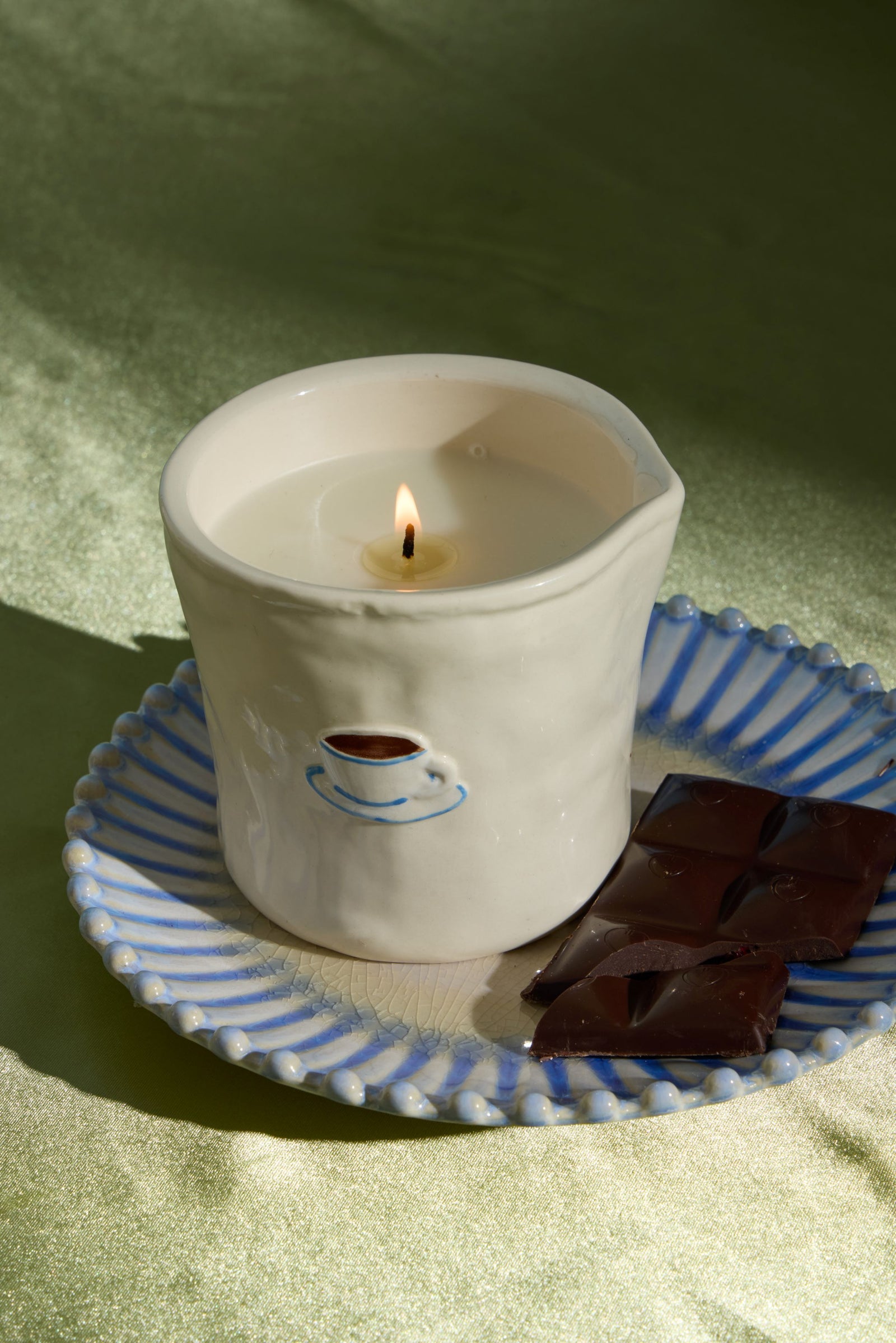 A lit white ceramic candle holder shaped like a small cup, placed on a blue-and-white striped saucer with dark chocolate pieces beside it, set against a softly lit olive-green fabric background.