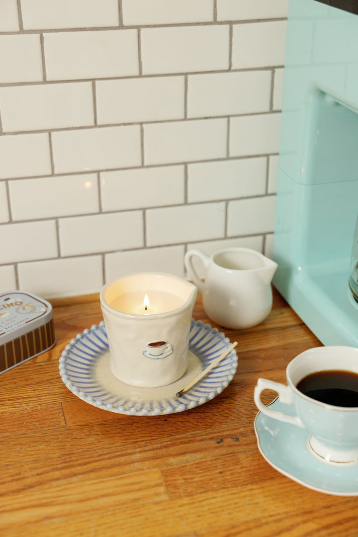 A lit cream-colored candle in a ceramic cup with a blue and white striped saucer, set on a wooden countertop next to a cup of coffee, a small white pitcher, and a white subway tile backsplash.