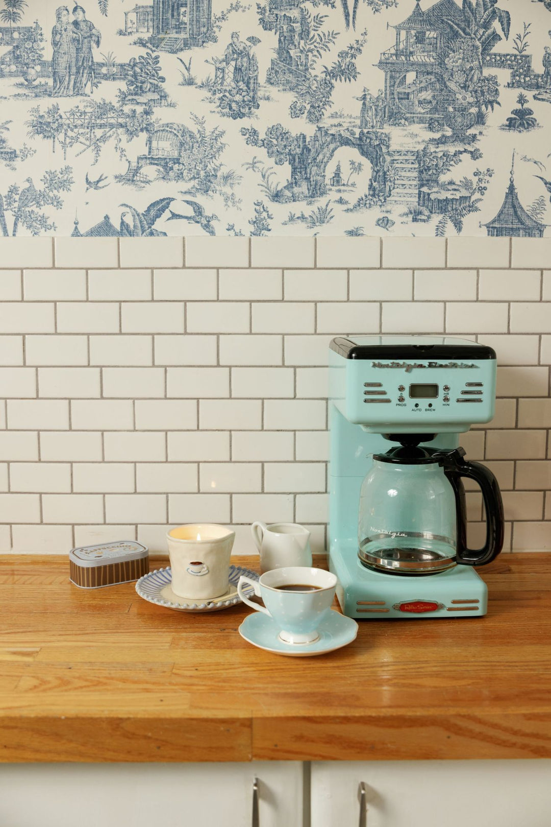 A retro-style mint green Hamilton coffee maker sits on a wooden countertop next to two cups and a small container, set against a white subway tile wall and a blue toile patterned upper wall.