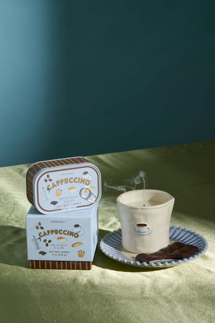 A coffee-themed scented candle set on a sage green fabric surface against a dark teal background. The set includes a square tin and matching box labeled &