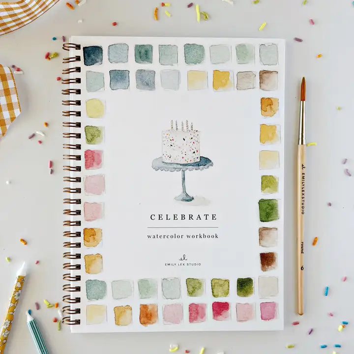 An Emily Lex watercolour workbook with a Celebration theme on a white background with sprinkles and a paintbrush next to it. 