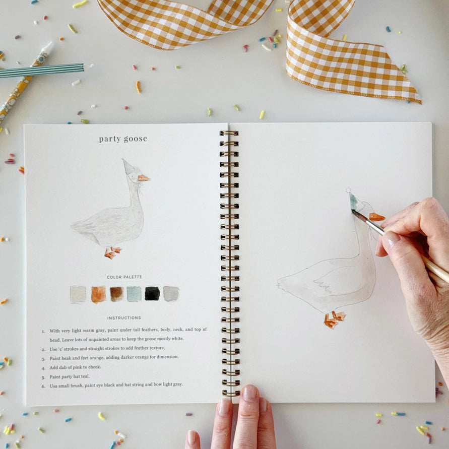 A hand holding a paintbrush sketches a goose on an open spiral notebook, with printed goose artwork, color swatches, and instructions visible on the left page. The scene is styled on a light surface with confetti, a gingham ribbon, and a striped paintbrush.