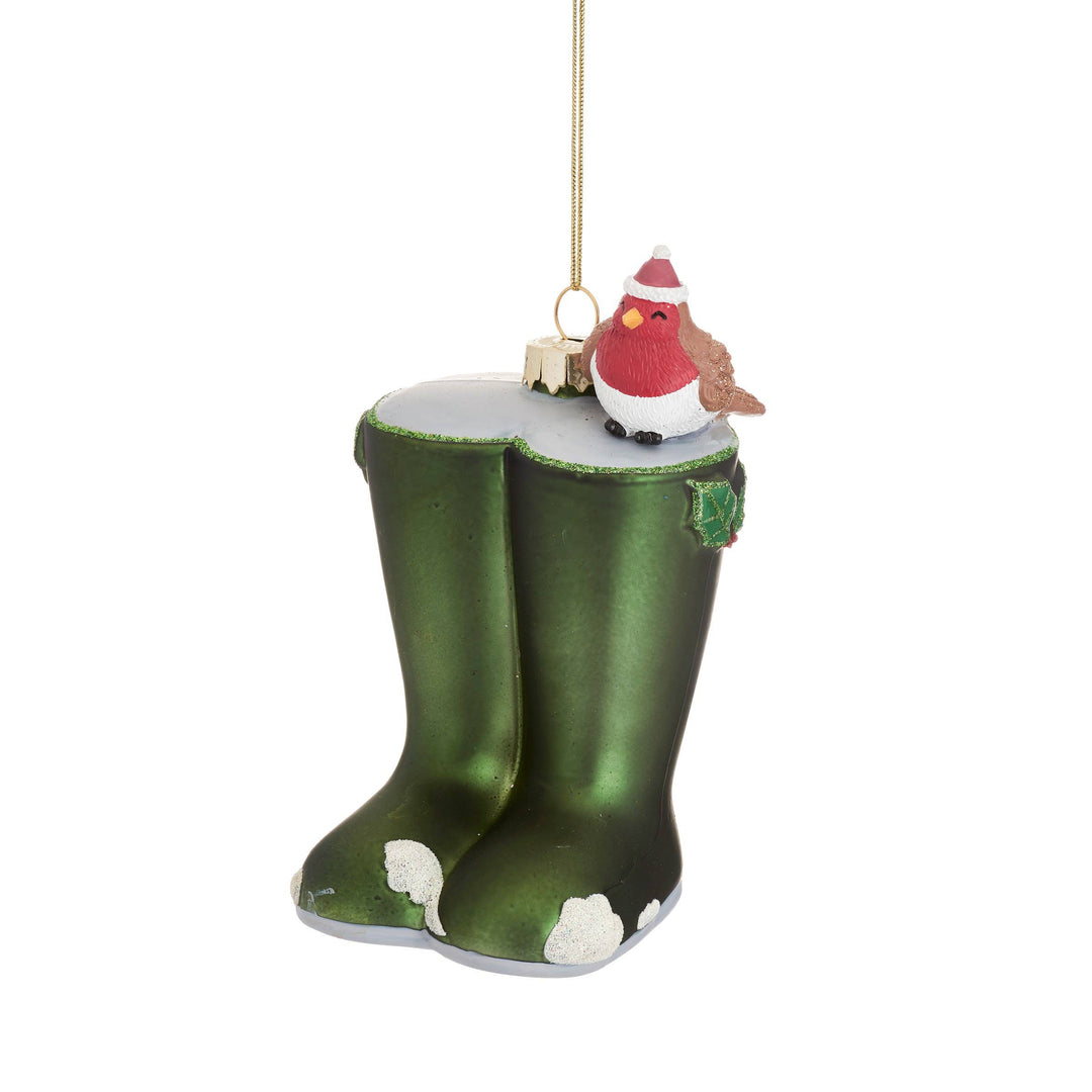 A Christmas ornament shaped like a pair of green rain boots with white snow patches, topped with a small red bird wearing a Santa hat, hanging from a gold string on a white background.