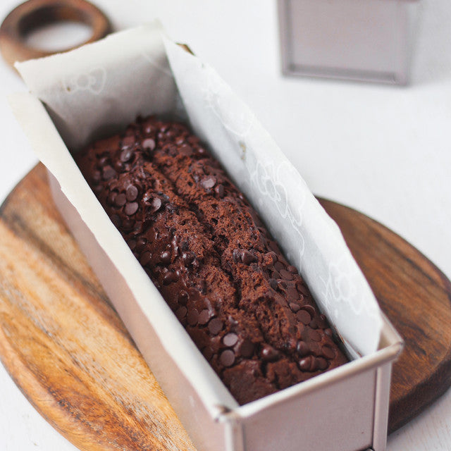 Loaf of chocolate bread with chocolate chips in a white loaf pan on a wooden surface.