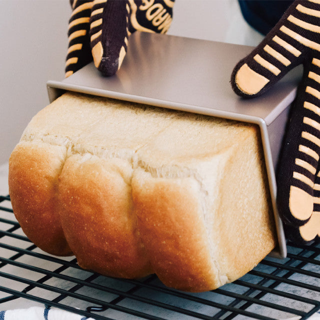 Loaf of bread being removed from a metal loaf pan with oven mitts on a cooling rack.