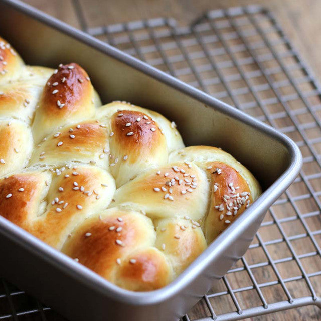 Baked bread rolls with sesame seeds in a metal loaf pan on a cooling rack.