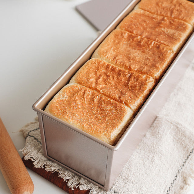 Loaf of bread in a metal loaf pan on a white surface with a wooden rolling pin.
