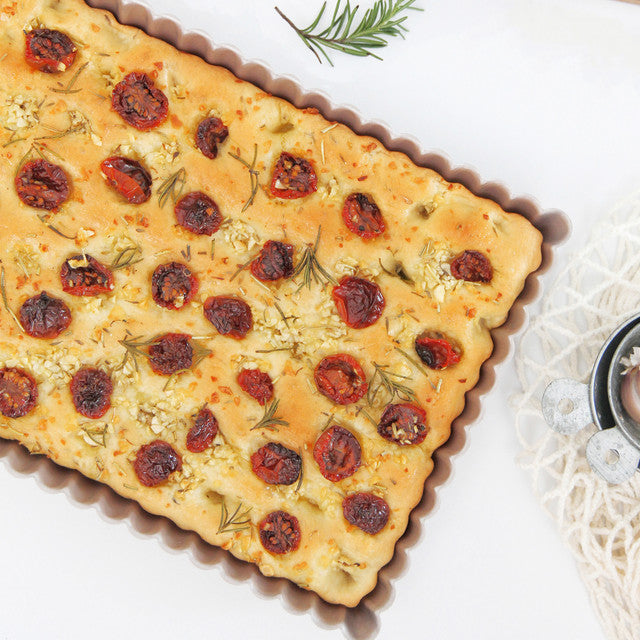 Rectangular focaccia bread with dried tomatoes and rosemary on a white surface.