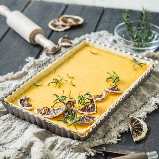 Rectangular tart with a yellow filling, garnished with dried lemon slices and rosemary, on a wooden surface.