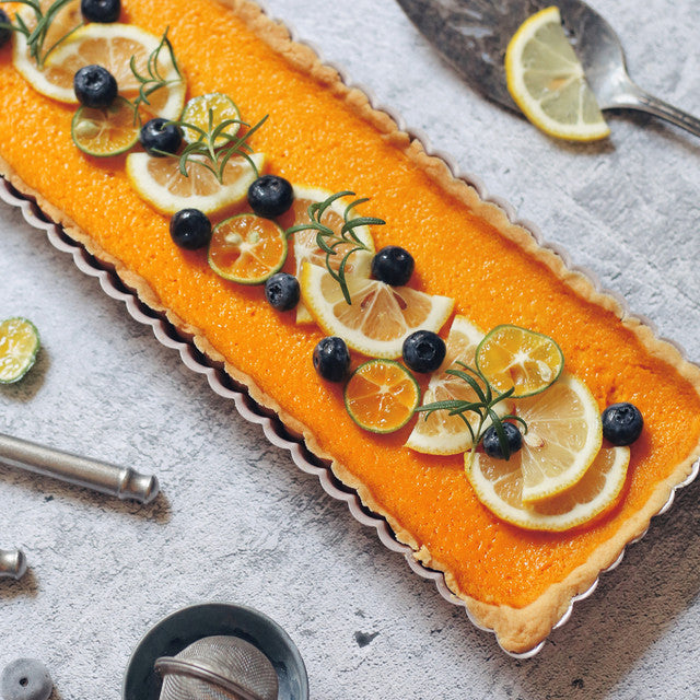 Rectangular tart with orange filling, lemon slices, and blueberries on a textured surface.