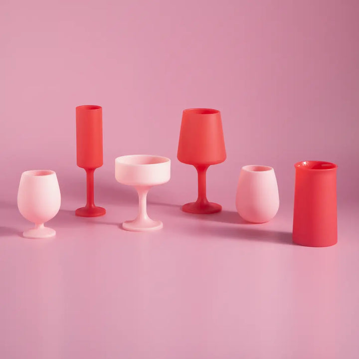 Six matte silicone drinking glasses in various shapes and colors of pink and red, arranged on a matching pink background.