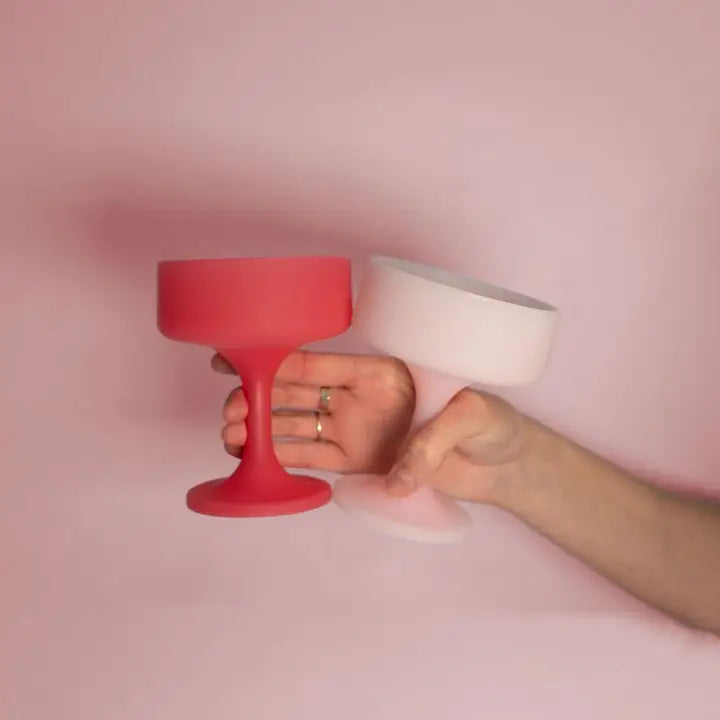 A hand holds two matte silicone cocktail cups against a solid pink background, one in coral red and the other in pale pink.